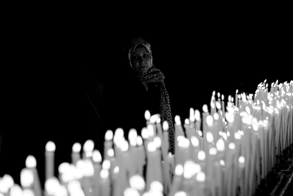 Black and white photo shows tightly packed rows of candles in the foreground. An older woman wearing a scarf on her head and around her neck is barely visible in the background.